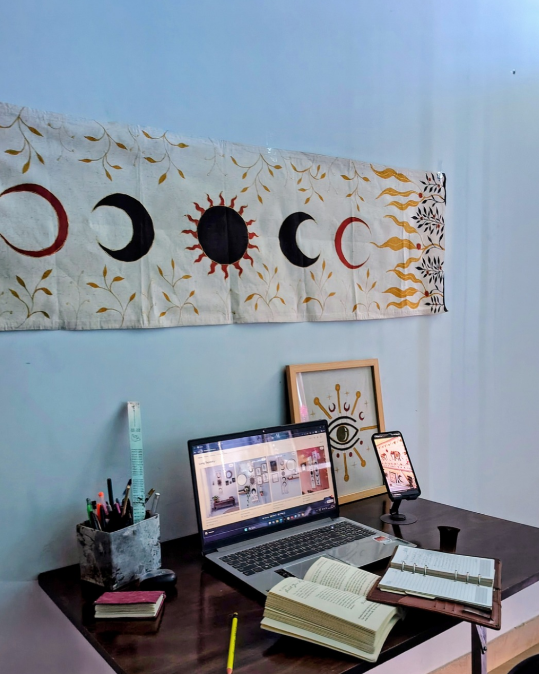 Desk setup with laptop, books, and decor items against a wall with a celestial tapestry. sun and moon and leaves, hand painted room decor item, good for gifting purpose.