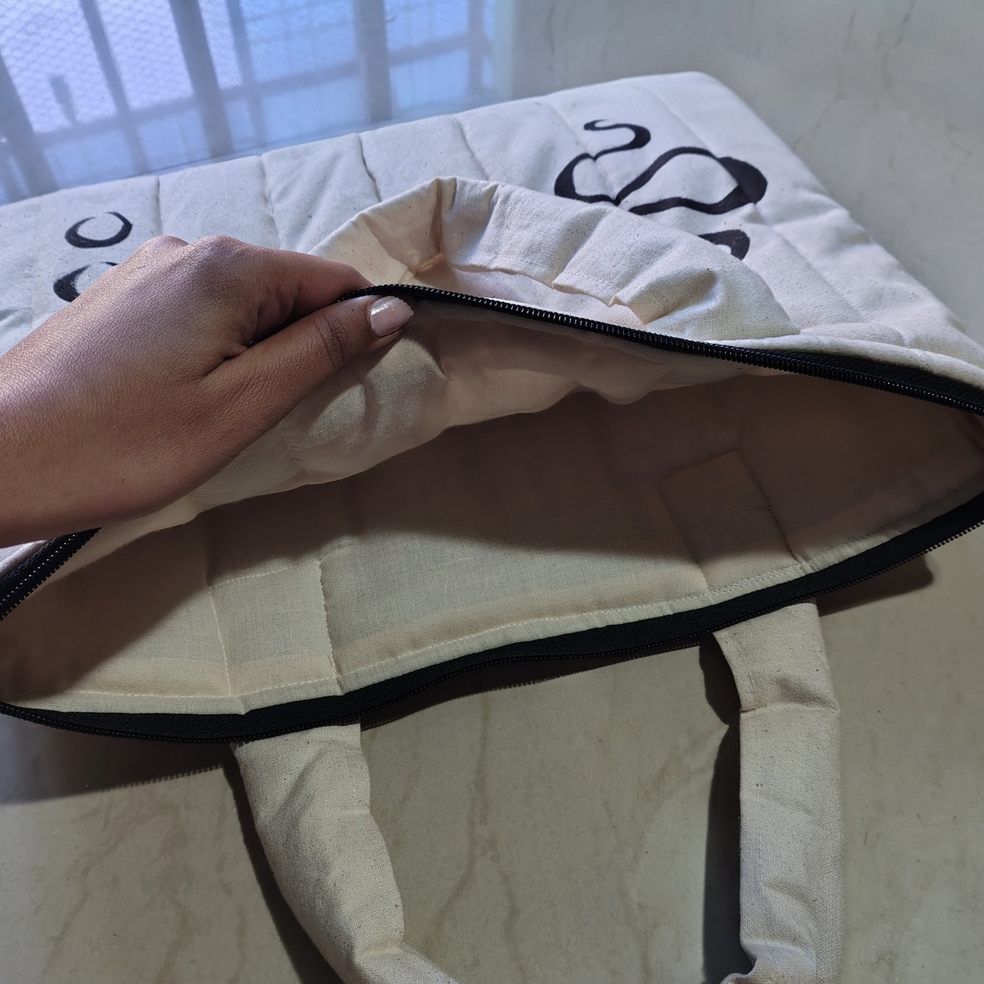 Person opening a beige bag with a black zipper on a light-colored surface. puffy laptop bag, hand painted snake, 