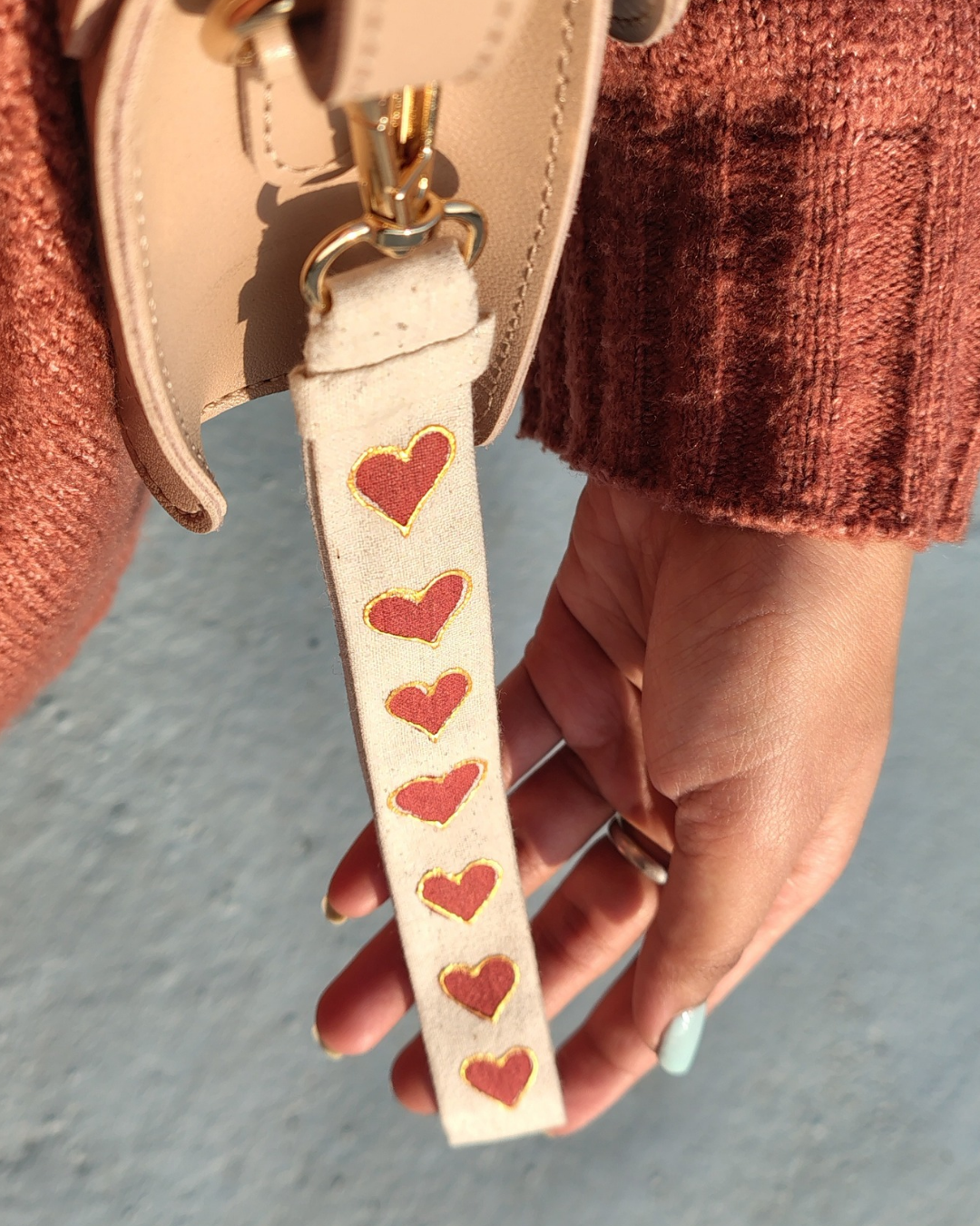 Hand holding a beige strap with red heart patterns, wearing a pink textured sweater. bag accessory, key chain