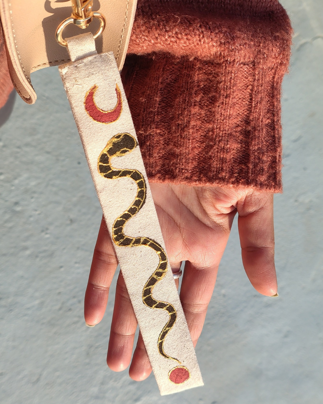 Strap with snake design held by a hand against a neutral background. bag accessory, key chain.